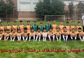عکس گروهی تیم فوتبال کودکان با لباس‌های زرد و مشکی در زمین چمن، همراه با مربیان ایستاده و بازیکنان نشسته، با پس‌زمینه‌ای از نرده‌های ورزشگاه و درختان