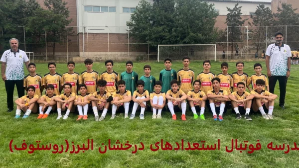 عکس گروهی تیم فوتبال کودکان با لباس‌های زرد و مشکی در زمین چمن، همراه با مربیان ایستاده و بازیکنان نشسته، با پس‌زمینه‌ای از نرده‌های ورزشگاه و درختان