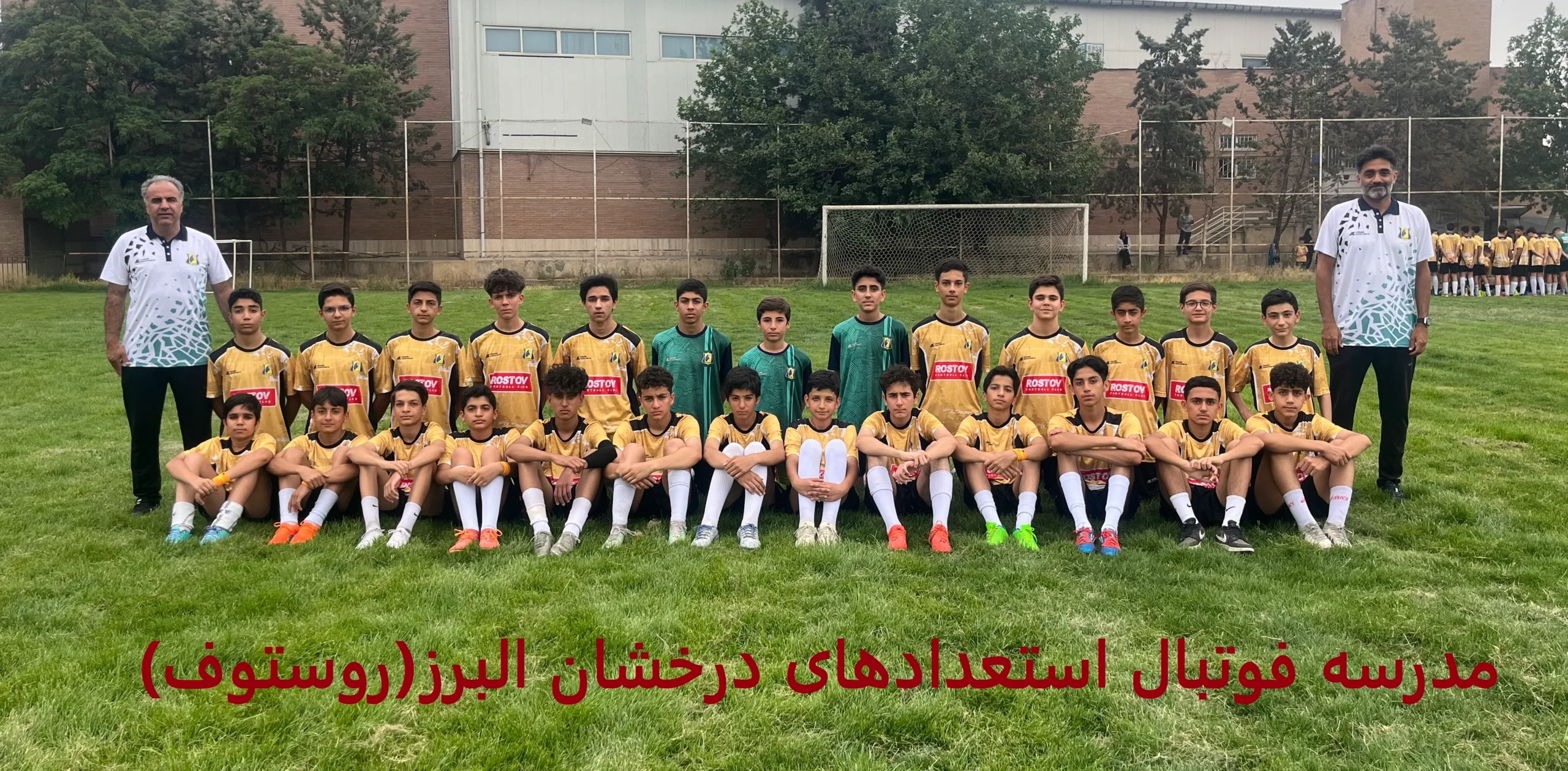 عکس گروهی تیم فوتبال کودکان با لباس‌های زرد و مشکی در زمین چمن، همراه با مربیان ایستاده و بازیکنان نشسته، با پس‌زمینه‌ای از نرده‌های ورزشگاه و درختان